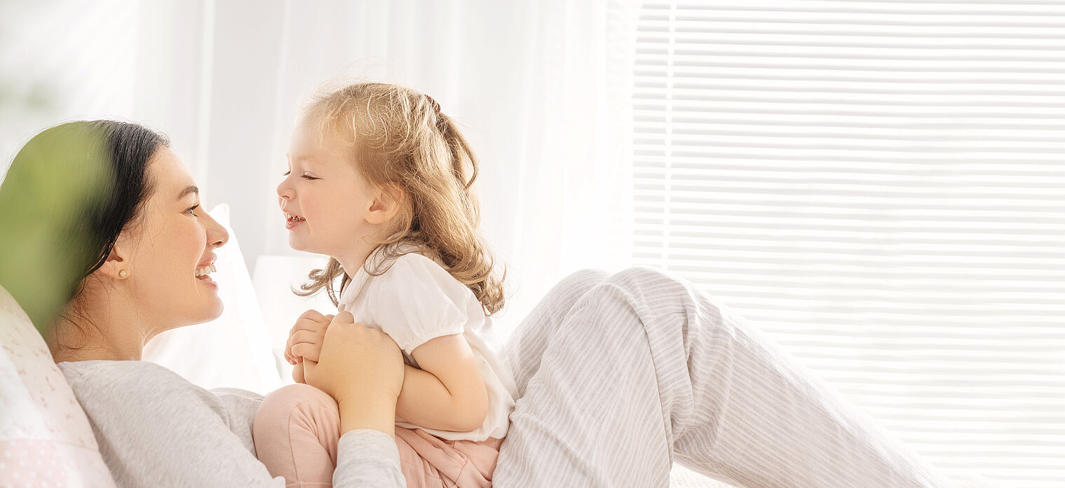 Mother and young daughter smiling at each other while lying down in a bright room with light filtering through the blinds.