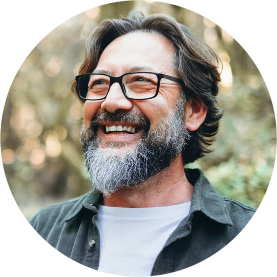 A smiling man with a beard and glasses standing outdoors, looking up.