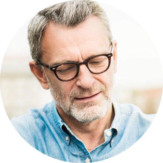 A close-up of an older man wearing glasses and a denim shirt, looking content with a gentle smile on his face.