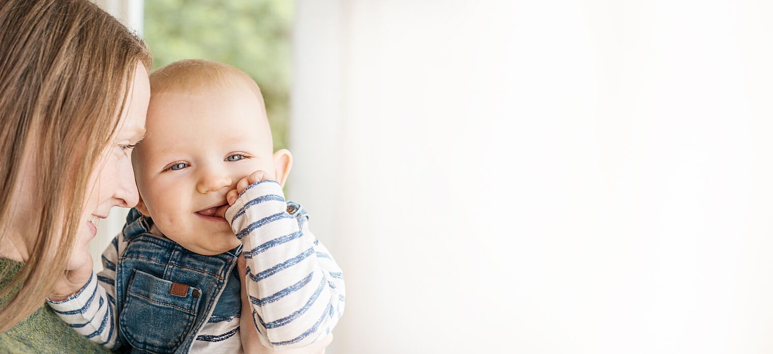 Mother nuzzling her baby, who is smiling and wearing a striped shirt and denim overalls, in a brightly lit room