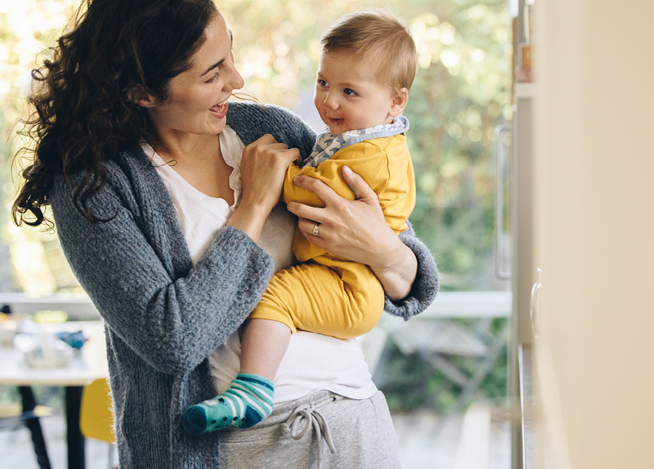 Mother smiling and holding her baby dressed in a yellow outfit, standing in a bright room with a window and greenery in the background.